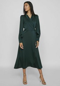Abito avvolgente in satin verde scuro con maniche a palloncino lunghe, vita sottolineata da una cintura e una silhouette A-line fluida. Indossato con tacchi dorati con cinturini.