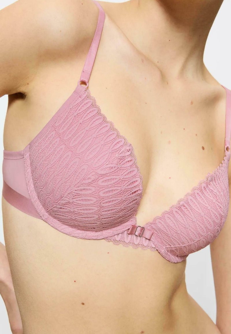 Jasnoróżowy koronkowy stanik z teksturowanym wzorem, wsparciem w postaci fiszbin, regulowanymi ramiączkami oraz małą kokardką na środku przodu.