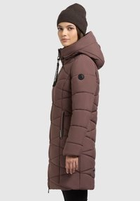 Polsterdatud pruun tikitud jakk kapuutsiga, millel on lukuga taskud ja logo aktsendid, näidatud küljelt tume kudumine beanie.