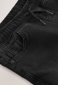 Close-up af sorte denim joggerbukser med elastisk talje, snøre, frontlomme og syningsdetaljer.