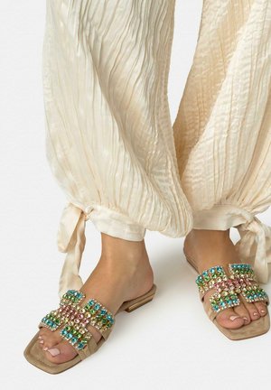 Pies con sandalias beige adornadas con piedras preciosas multicolores, vistiendo pantalones crema sueltos con textura, atados en los tobillos, sobre un fondo blanco.