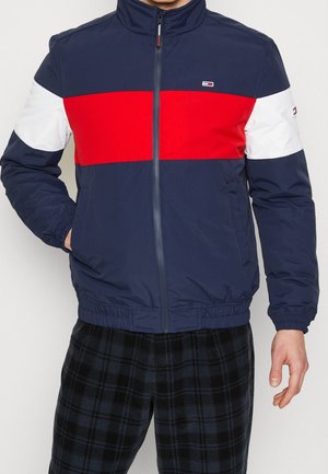Hombre vestido con una chaqueta azul marino con rayas horizontales rojas y blancas y pantalones oscuros de cuadros, con las manos en los bolsillos.