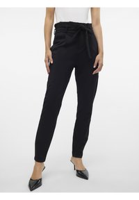 Pantalon noir taille haute avec une large ceinture nouée, jambes fuselées et poches latérales. Texture lisse et coupe ajustée, associé à des escarpins noirs à bouts pointus.