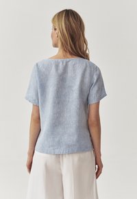 Blonde vrouw in een lichtblauwe blouse met korte mouwen en witte wijde broek, met haar rug naar de camera gekeerd tegen een eenvoudige witte achtergrond.