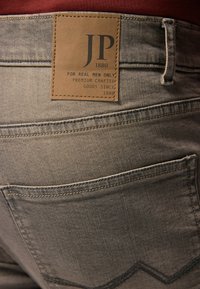 I jeans di denim grigio presentano una toppa in pelle marrone con testo in rilievo e due tasche posteriori con cuciture curve. Il tessuto ha una texture leggermente usurata.