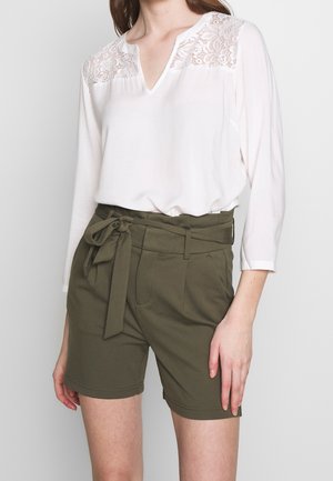 Kvinde iført hvid bluse med lange ærmer med blondeindsats og olivengrønne højtaljede shorts bundet med stofbælte i taljen.