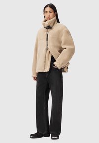 Beżowa bluza polarowa z wysokim kołnierzem i zamkiem, z czarnymi akcentami. Nosi na sobie luźne czarne spodnie i czarne obuwie.