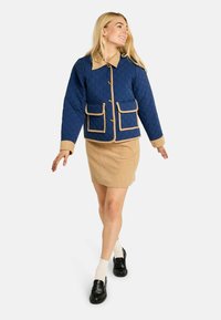 Jachetă matlasată bleumarin cu accente bej, buzunare frontale mari și guler; combinată cu o fustă de catifea și mocasini negri.