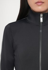 Čierna bunda na zips s vysokým golierom, vyrobená z hladkej tkaniny. Má strieborný zips a sivé akcenty pozdĺž ramien.