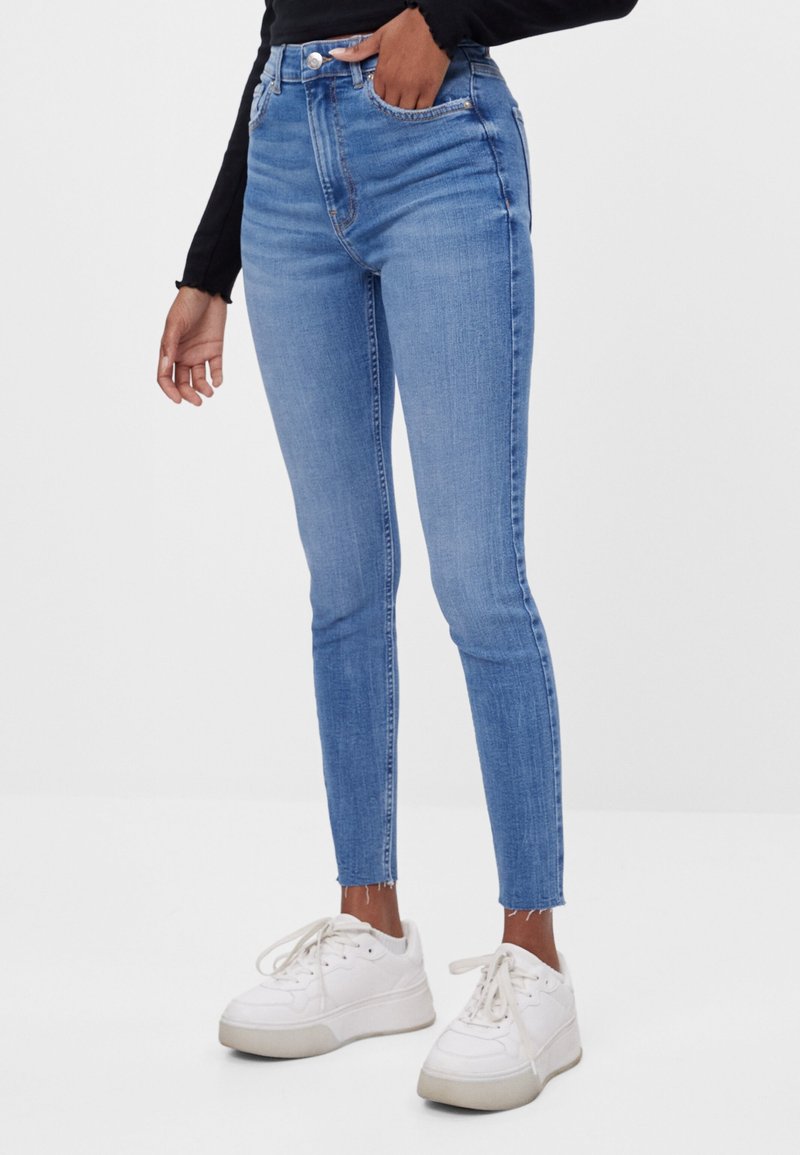 Jeans de talle alto en azul claro con una forma ajustada, un ligero desgaste y dobladillos sin terminar, combinados con zapatillas blancas de suela gruesa.