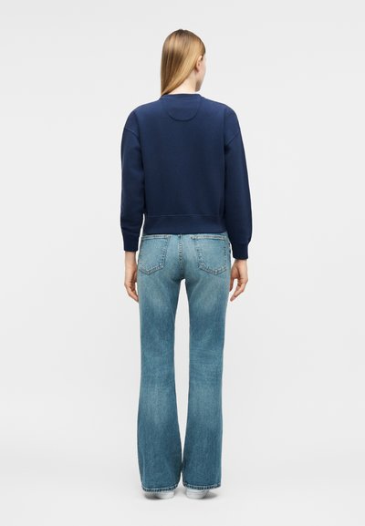 Γυναίκα με μακριά ξανθά μαλλιά που φοράει μπλε σκούρο φούτερ και μπλε ψηλόμεσα τζιν καμπάνα, να στέκεται με την πλάτη προς τα εμπρός σε απλό φόντο.