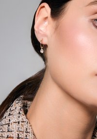 Złoty kolczyk w formie obręczy z przezroczystym, gruszkowatym zawieszką wisi z płatka ucha, eksponowany na tle teksturowanej tkaniny.