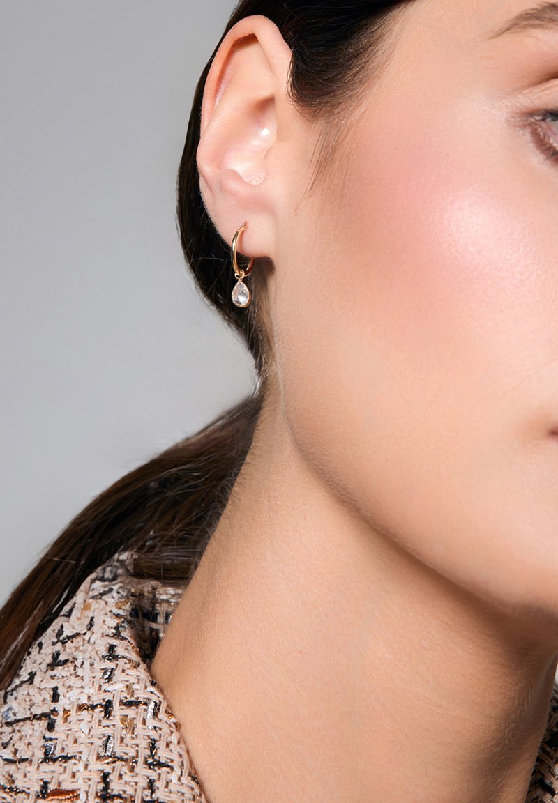 Złoty kolczyk w formie obręczy z przezroczystym, gruszkowatym zawieszką wisi z płatka ucha, eksponowany na tle teksturowanej tkaniny.