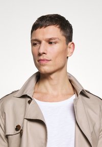 Beige Trenchcoat mit großen braunen Knöpfen, strukturiertem Kragen und weicher Textur, getragen über einem weißen Hemd. Gepflegtes, maßgeschneidertes Design.