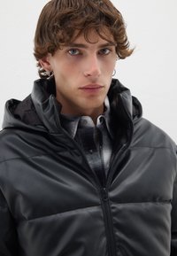 Chaqueta negra acolchada con capucha, con una textura sintética suave, cierre de cremallera y una camisa de cuadros gris en contraste debajo.