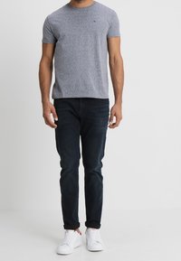 Šedé tričko s krátkým rukávem v kombinaci s tmavě modrými slim-fit džíny. Model nosí bílé tenisky s červenými akcenty. Jednoduchý, ležérní outfit.