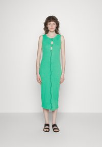 Vestido midi verde sem mangas com botões à frente, tecido texturizado e bordas recortadas; combinado com sandálias pretas.