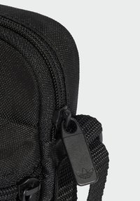 Sacca in tessuto nero con superficie strutturata, dotata di una zip ben visibile e un'etichetta con un logo. Include una tracolla intrecciata per il trasporto.