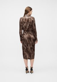 Γυναίκα που στέκεται με την πλάτη γυρισμένη, φοράει ένα καφέ φόρεμα με σχέδια και μακριά μανίκια μέχρι το μέσο της γάμπας και μαύρα πέδιλα με ψηλό τακούνι, σε λευκό φόντο.