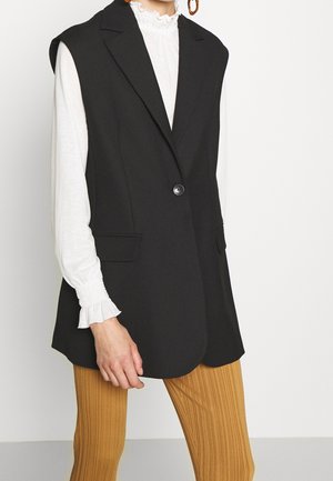 Woman wearing black sleeveless blazer over white long-sleeve blouse with ruffled cuffs, paired with mustard ribbed pants.