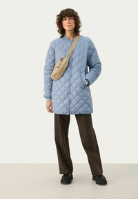 Lysblå quiltet lang jakke med ståkrave, med knaplukning og sidelommer. Sammen med brune bukser og en beige crossbody taske.