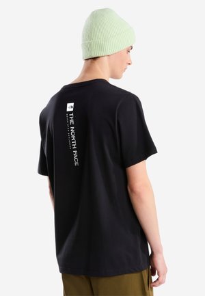 Persona joven que lleva una camiseta negra de The North Face con un logotipo blanco vertical en la espalda y un gorro de punto verde claro.