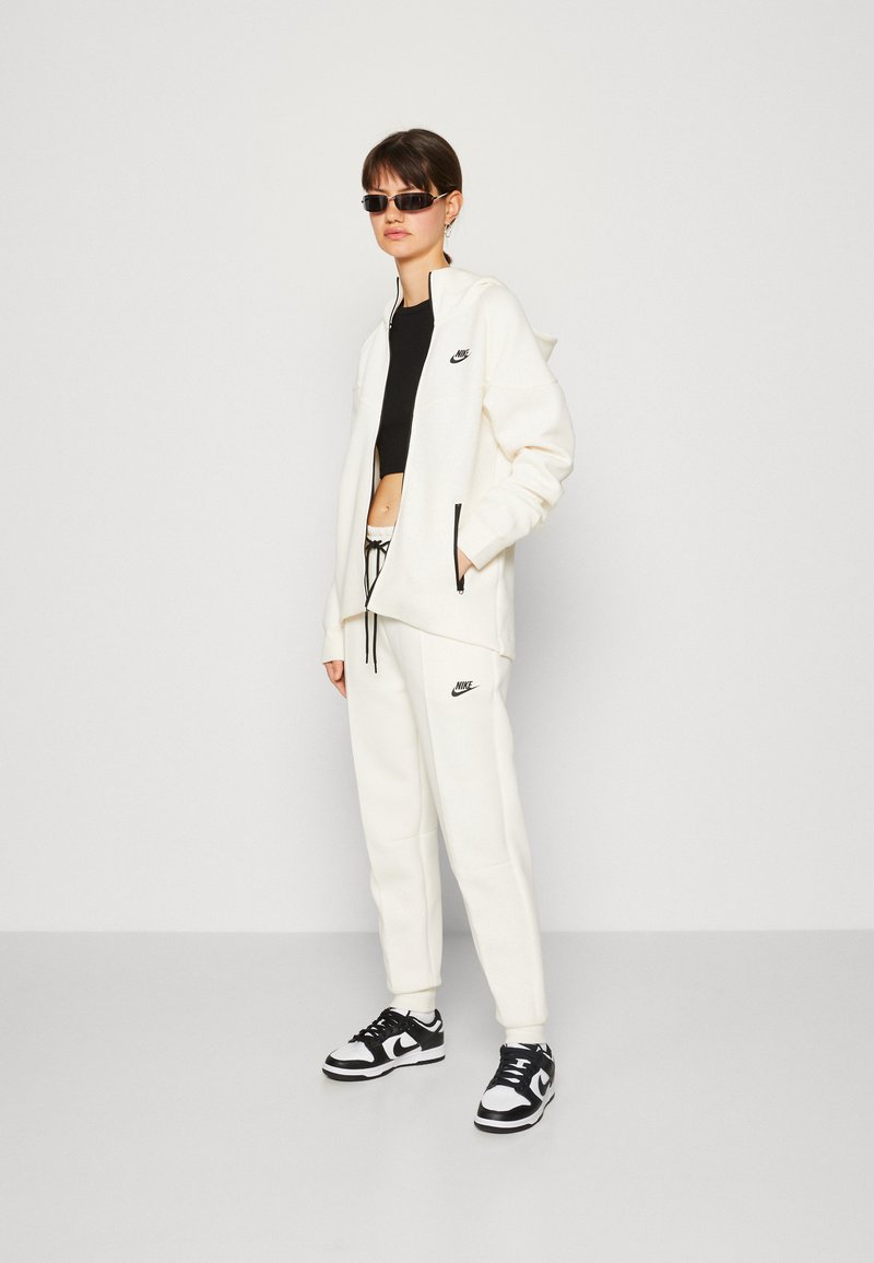 Sudadera blanca con cremallera y joggers a juego, con acentos de logo negro, combinados con zapatillas en blanco y negro. La modelo lleva gafas de sol oscuras.