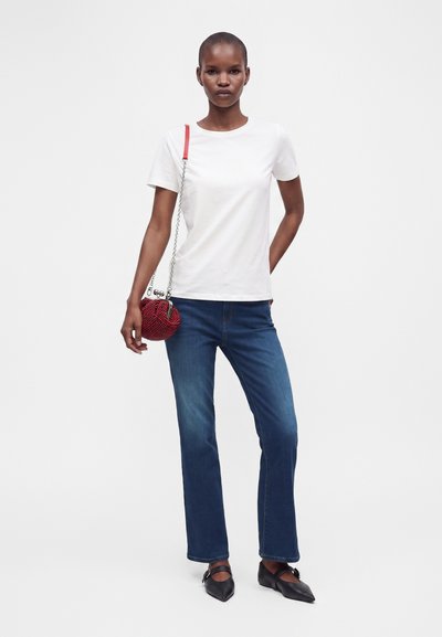 Femme portant un t-shirt blanc à manches courtes, un jean bleu, des ballerines noires pointues et tenant un petit sac rouge avec une chaîne.