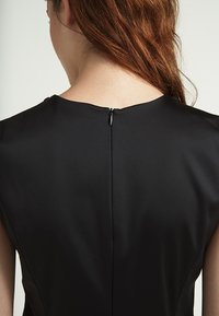 Schwarzes Kleid aus glattem, mattem Stoff. Verfügt über einen dezenten Ausschnitt und einen senkrechten Verschluss am Rücken mit einem kleinen Reißverschluss.