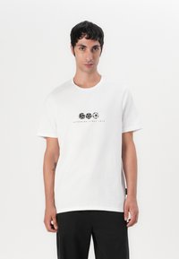 Vit bomulls t-shirt med korta ärmar, rund hals, med svart grafiskt tryck av sportbollar och texten "REPPING SINCE 1974."