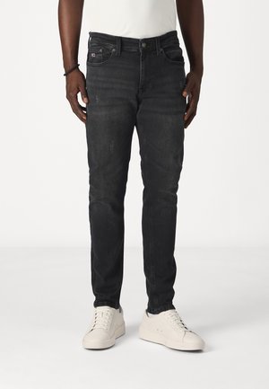 Sorte slim-fit jeans med let slid, båret af en person med hvide sneakers og en hvid skjorte, stående foran en ensfarvet baggrund.