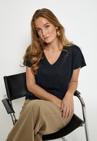 Maglietta blu navy con scollo a V e texture scintillante, maniche corte, abbinata a pantaloni beige chiaro, con una modella seduta su una sedia nera.