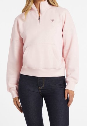 Kvinde iført lyserød kvart-lyntrøje med kængurulomme og mørkeblå tætsiddende jeans mod en ensfarvet hvid baggrund.