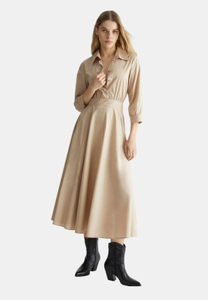 Abito midi beige a maniche lunghe con vita aderente, colletto, bottoni frontali e gonna voluminosa. Indossato con stivaletti neri con tacco.