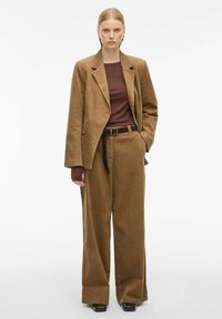 Blazer en velours côtelé marron et pantalon large assorti, associé à un haut ajusté bordeaux ; présente une ceinture et une texture lisse sur un fond neutre.