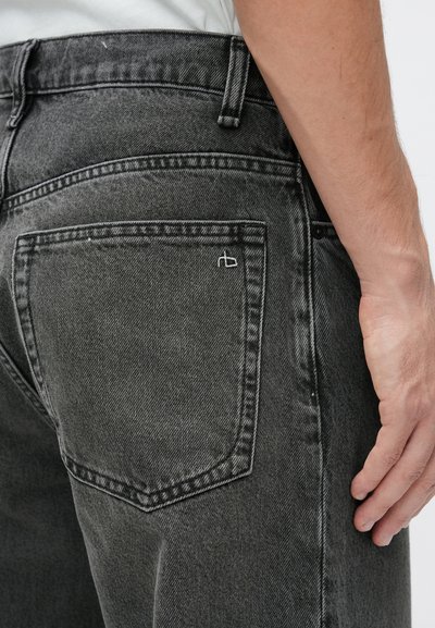 Nærbillede af en person iført slidte sorte denimjeans, der viser baglommen og en del af en hvid skjorte, der er stoppet ind.