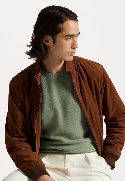 Jachetă bomber din suede maro, cu manșete striate, purtată peste un pulover texturat de culoare verde măslinie, asortată cu pantaloni de culori deschise.