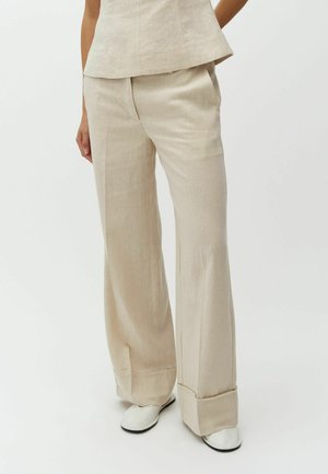 Pantaloni beige a gamba larga con risvolti indossati con scarpe basse bianche e una canotta beige abbinata su una persona in piedi su uno sfondo bianco.