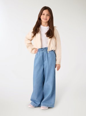 Ragazza sorridente con lunghi capelli castani che indossa una giacca beige chiaro, una camicia bianca, jeans larghi blu e sneakers rosa, in piedi con una mano sul fianco.