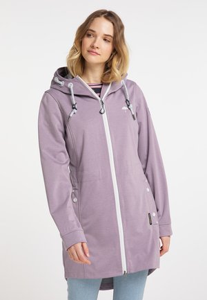 Jonge vrouw draagt een lange lavendelkleurige jas met capuchon, witte rits en koordjes, gecombineerd met lichtblauwe spijkerbroek.