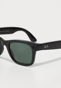 Occhiali da sole Ray-Ban con montatura in plastica nera, lenti verde scuro e una caratteristica della fotocamera sul lato. Logo visibile sulla tempia.