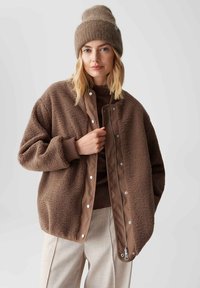 Braune Fleece-Bomberjacke mit satinartigem Innenfutter, gerippten Bündchen und Druckknopfverschluss; getragen über einem dunkelbraunen Oberteil und hellbraunen Hosen.