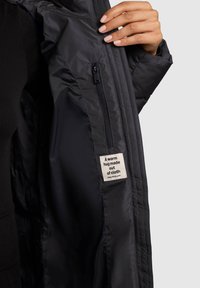 Čierna izolovaná bunda s vnútorným vreckom na zips; obsahuje textilnú štítok s nápisom "Teplé objatie vyrobené z látky." Jemná, hladká textúra.