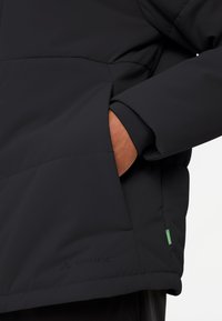Zwarte jas met een gewatteerd ontwerp, elastische manchetten en een zijzak. "Vaude"-logo zeefdruk op de onderzijde van de voorkant. Groene accentdetails zichtbaar.