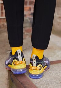 Nike VaporMax Sneaker mit einem mehrfarbigen gestrickten Obermaterial, gelben Akzenten, transparenter Sohle und "LOCK"-Tags auf der Rückseite. Schwarze Hose und gelbe Socken.