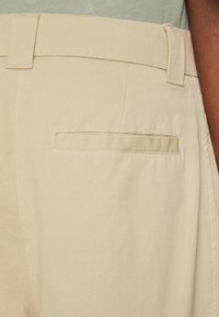 Beige színű pamut nadrág lapos elülső résszel, egy hátsó zsebbel és kontrasztos varrással. Az anyag sima, enyhén texturált felülettel rendelkezik.