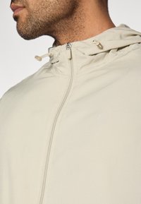 Világos bézs színű cipzáras kapucnis dzseki, sima textúrával, finom logóval és állítható húzózsinórokkal a nyaknál.