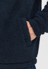 Veste en polaire bleu marine avec un tissu texturé et duveteux. Elle possède une poche latérale à l'avant, avec une coupe décontractée et des manches à poignets.
