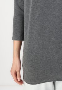 Top grigio, leggero, con maniche a tre quarti, texture morbida, taglio dritto e orlo arrotondato. Presenta un design semplice e minimalista.