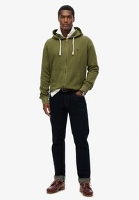 Sudadera con cremallera de color verde oliva con cordones blancos, dos bolsillos frontales, llevada sobre una camiseta blanca, combinada con jeans oscuros y zapatos marrones.